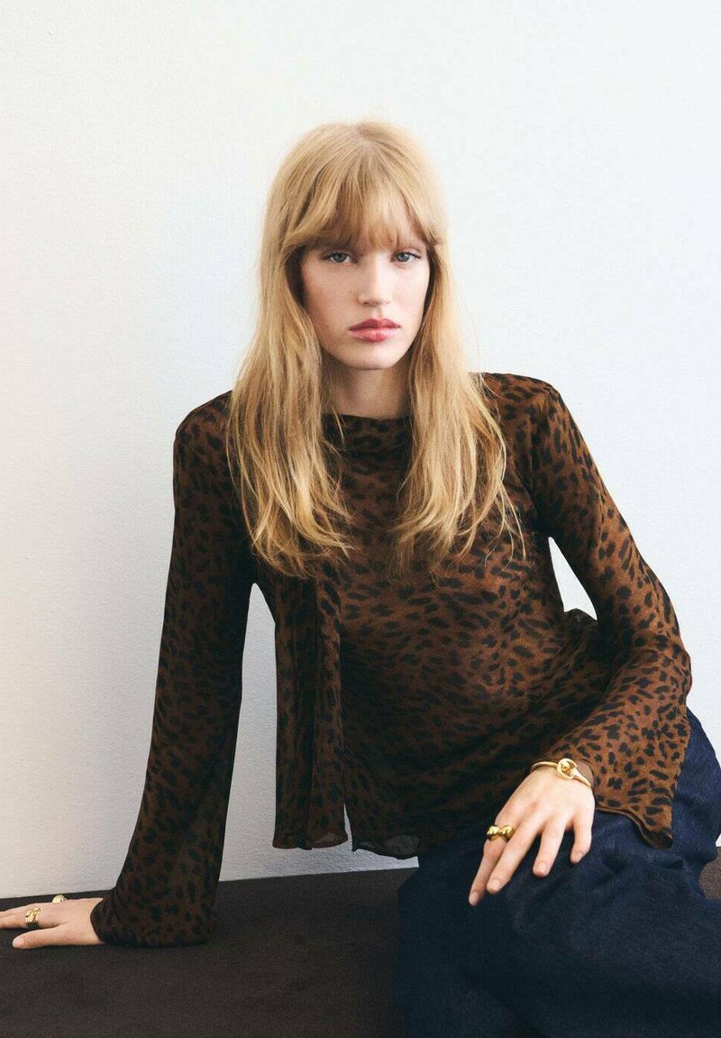 Braune durchsichtige Bluse mit schwarzem Leopardenmuster, mit langen Ärmeln und fließendem Design. Getragen mit Jeans und goldenen Schmuckakzenten.