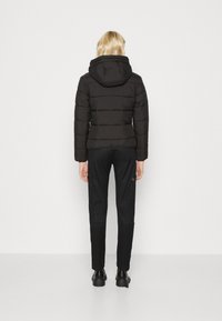 Doudoune noire avec des coutures horizontales, capuche ajustée et manches longues, associée à un pantalon noir et des chaussures, vue de dos.