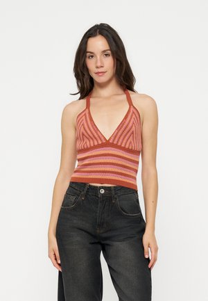 Femme aux longs cheveux foncés portant un crop top rayé rose et orange à bretelles et un jean noir taille haute sur fond blanc.