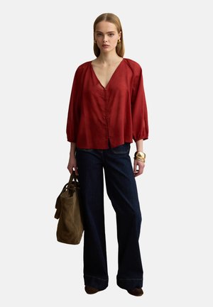 Vrouw draagt een losse rode V-halsblouse, donkere wijde jeans, gouden armbanden en houdt een grote olijfgroene handtas vast.