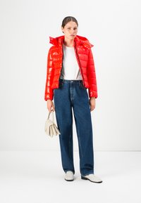Vrouw in een felrode pufferjack, witte shirt, losse donkerblauwe jeans, witte loafers, die een beige handtas vasthoudt tegen een witte achtergrond.