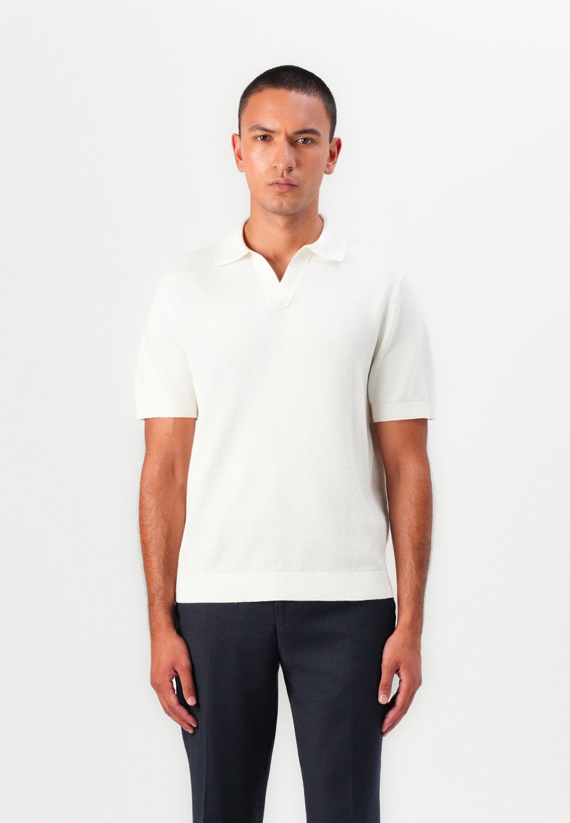 Weißes Poloshirt mit kurzen Ärmeln aus strukturiertem Strick, mit Kragen und geripptem Saum. Kombiniert mit dunklen Hosen, vor einem neutralen Hintergrund.