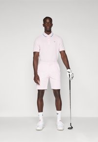 Polo de cor rosa claro com colarinho azul, acompanhado de calções a condizer. Ténis brancos e um taco de golfe completam o conjunto desportivo.