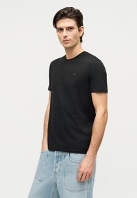 Νέος άνδρας με σκούρα μαλλιά, φορώντας μαύρη κοντομάνικη μπλούζα και ανοιχτό μπλε τζιν, στέκεται μπροστά σε ένα απλό φωτεινό φόντο.