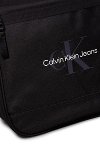 Fekete szövet üzenetküldő táska sima textúrával, a "Calvin Klein Jeans" felirattal és egy hangsúlyos "CK" díszítő elemmel.