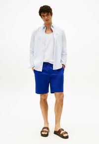 Camicia button-up a righe azzurro chiaro, canotta bianca, pantaloni corti blu brillante e sandali marroni con doppie cinghie. Outfit estivo casual.
