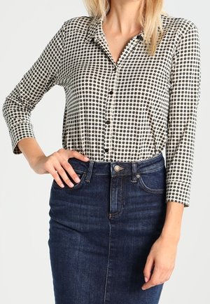 Kvinde iført en sort-hvid mønstret skjorte med knapper, stoppet ned i et mørkeblåt denimskørt, med hånden hvilende på hoften.