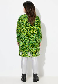 Abito verde a stampa leopardata con maniche lunghe, taglio allungato e orlo asimmetrico. Leggings argento e stivali neri alla caviglia completano l'outfit.