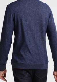 Uomo che indossa un maglione aderente a maniche lunghe di colore blu scuro e jeans neri, visto da dietro su uno sfondo neutro.