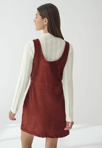 Rostfarbendes Cordkleid mit einem runden Rückenausschnitt, kombiniert mit einem strukturierten weißen Oberteil mit langen Ärmeln und horizontalen Streifen.