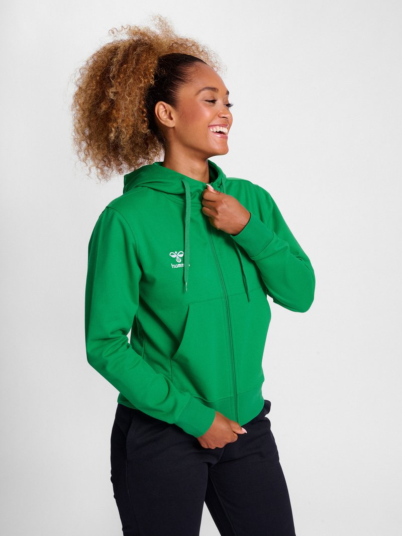 Frau mit lockigem Haar in einem grünen Kapuzenpullover mit Reißverschluss und schwarzen Hosen, die lächelnd den Kragen des Hoodies vor einem schlichten weißen Hintergrund richtet.