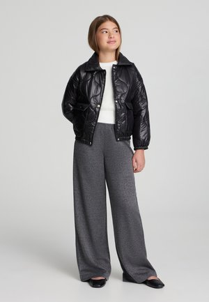 Giacca a vento nera con finitura lucida, design trapuntato e colletto. Abbinata a pantaloni grigi a gamba larga con texture e scarpe basse nere.