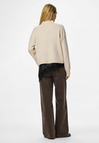 Bežani pleteni pulover s spuštenim ramenima, rebrastim manžetama i skraćenim rubom, složen preko crne majice s čipkastim detaljima, u kombinaciji s smeškim hlačama širokih nogavica.