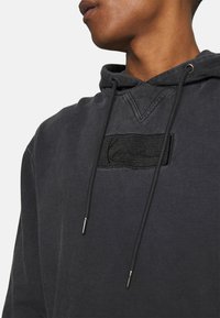 Sudadera con capucha de algodón gris oscuro, con un parche texturizado negro con letras grabadas, cordones y un cuello en forma de V acanalado.