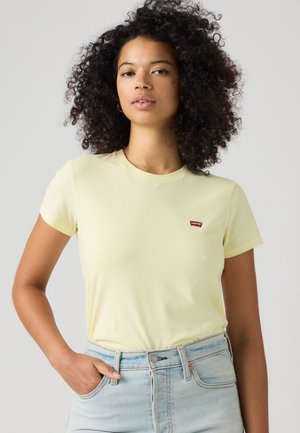 Mujer joven con cabello rizado que lleva una camiseta Levi's de color amarillo pálido y vaqueros azul claro, de pie con una mano en el bolsillo.