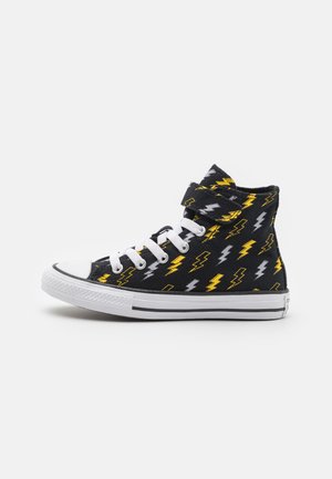 Zapatilla alta negra con patrón de relámpago en amarillo y gris, cordones blancos, puntera de goma y suela blanca con franja negra.
