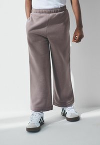 Gai brūni sporta bikses ar elastīgu jostasvietu un sānu kabatām, izgatavotas no mīksta materiāla. Valkātas ar baltām sporta kurpēm ar melniem akcentiem.