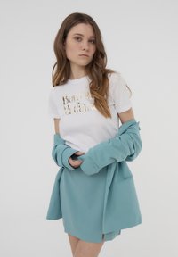 Maglietta bianca con testo in foil dorato, indossata sotto una giacca leggera color verde acqua. La giacca ha una vestibilità comoda e tasche.