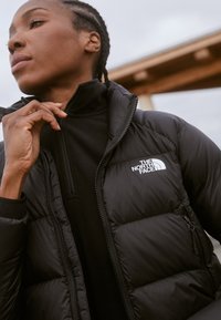 Giacca a vento nera con design trapuntato, dotata di chiusura con zip e logo sul lato sinistro del petto. Strato interno in fleece nero con colletto alto.