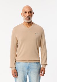 Beige v-ringad tröja i en mjuk stickad kvalitet, med ribbade manschetter och nederkant; accentuerad med en liten grön logotyp på bröstet. Bärs med ljusblå jeans.