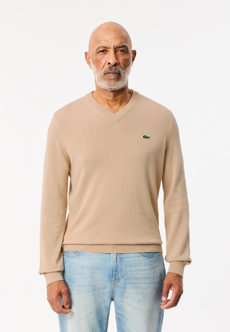 Beige v-ringad tröja i en mjuk stickad kvalitet, med ribbade manschetter och nederkant; accentuerad med en liten grön logotyp på bröstet. Bärs med ljusblå jeans.