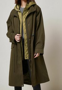 Manteau long vert olive avec une doublure matelassée verte olive, des boutons-pression et une capuche ajustable. Texture douce avec une coupe ample, présenté partiellement dézippé.