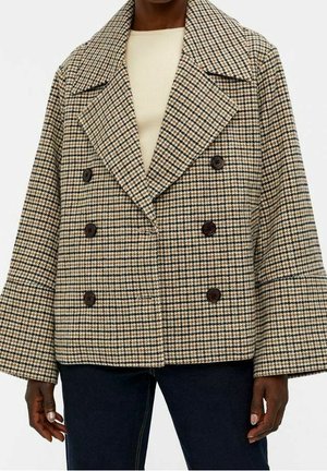 Dobbeltbrystet beige og sort hundetandsfrakke med brede revers, båret over en cremefarvet ribbet top og mørkeblå jeans.