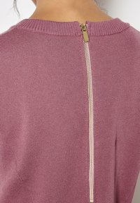 Maglione lavorato a maglia in rosa polveroso con scollatura a costine e un dettaglio in metallo dorato sulla cerniera centrale sul retro. Texture morbida e design aderente.