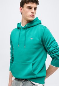 Νέος άνδρας που φοράει μπλε-πράσινη μπλούζα με κουκούλα, τσέπη μπροστά και κορδόνια, στέκεται με τα χέρια πίσω από την πλάτη, κοιτάζοντας προς τα πλάγια.