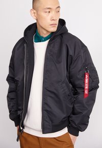Black bomber jacket with a zip front, ribbed cuffs, and hood; features utility patch with red "Remove before flight" tag.