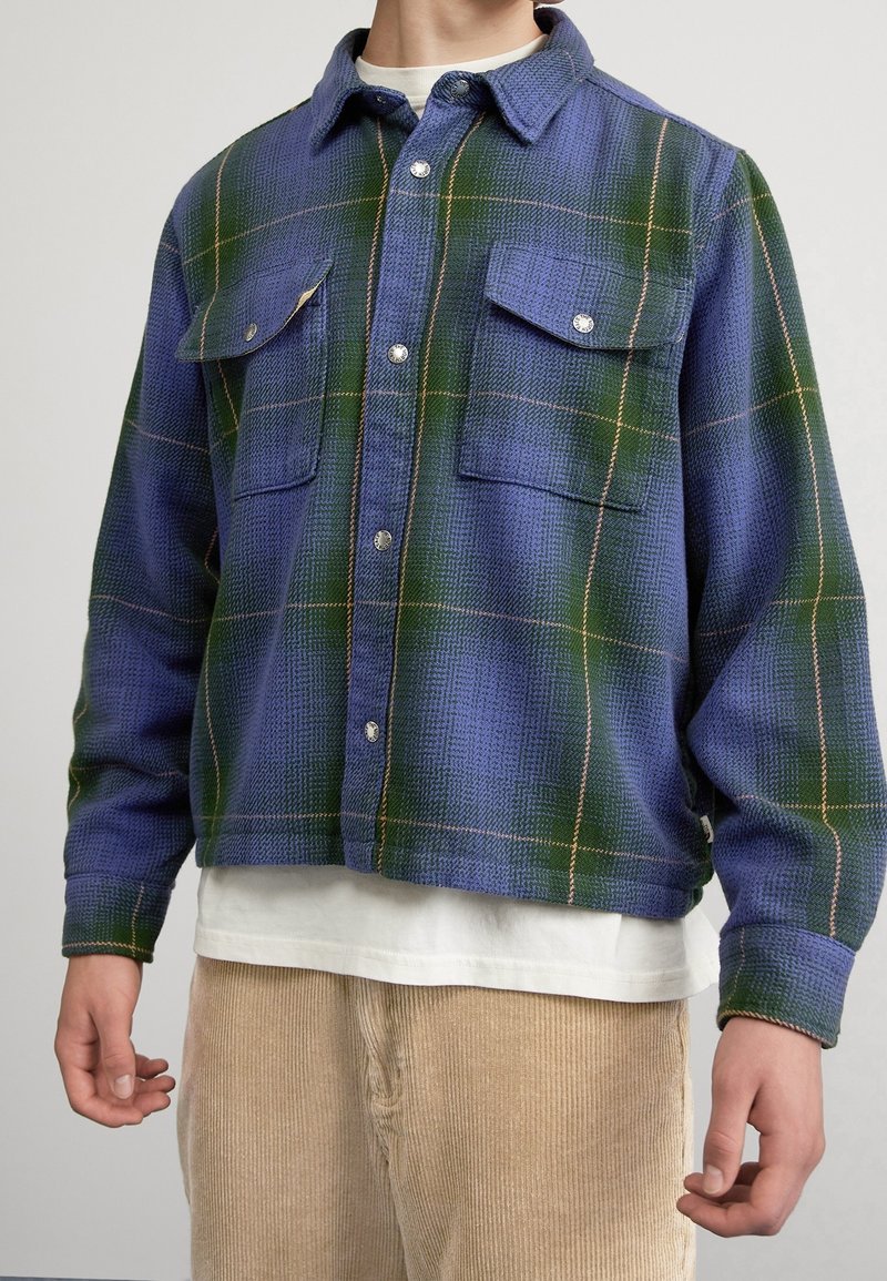Rutig flanellskjorta i blått och grönt med ljusbruna accenter, har två bröstfickor och silverknappar. Tyget har en strukturerad yta.