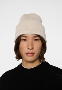 Beige stickad mössa med ribbad textur och uppvikt brätte. Subtil logodetalj framtill. Bärs tillsammans med en svart tröja.