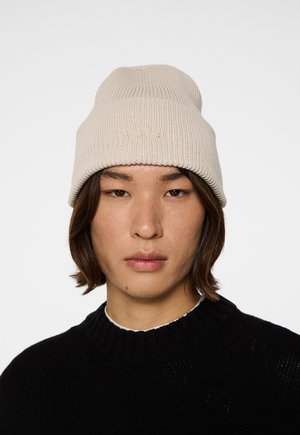 Beige gebreide beanie met ribbeltextuur en een omgeslagen rand. Subtiele logodetails aan de voorkant. Draagt het met een zwarte trui.