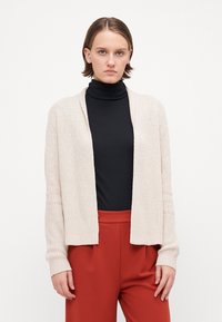 Beige strikket cardigan med åpen front, ribbet tekstur, lange ermer og avslappet passform, lagd over en svart genser med høy hals og røde bukser.