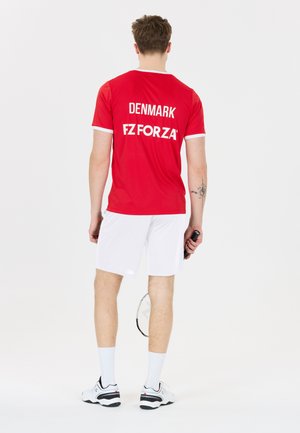 Mand, der holder en badmintonketcher, står med ryggen mod, iført en rød dansk sportstrøje og hvide shorts på en ensfarvet hvid baggrund.