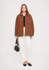 Giacca in pile marrone con chiusura a bottoni frontale, colletto e una tasca, abbinata a pantaloni bianchi a gamba larga e ballerine nere a punta.