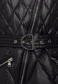 Musta tikattu takki, jossa kiiltävä pinta, vinottain kulkeva timanttikuvio, hopeanvärinen vetoketju ja vyötäröllä erinomainen metallisolki.