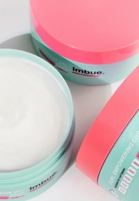 Twee potten haarproduct met mintgroene bases en roze deksels. Eén pot bevat witte gel, gelabeld als "Imbue Curl Empowering Cream."