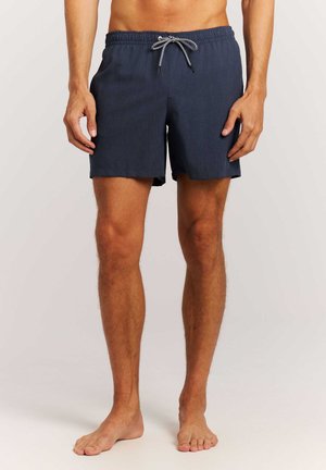 Homme portant un short de bain bleu marine avec un cordon noir et blanc, debout pieds nus devant un fond uni.