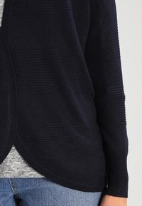 Laivastonsininen neuletakki, jossa on tekstuuriraitoja, auki oleva etuosa ja ribbikujat, yhdistettynä vaaleanharmaaseen toppiin ja sinisiin farkkuihin.