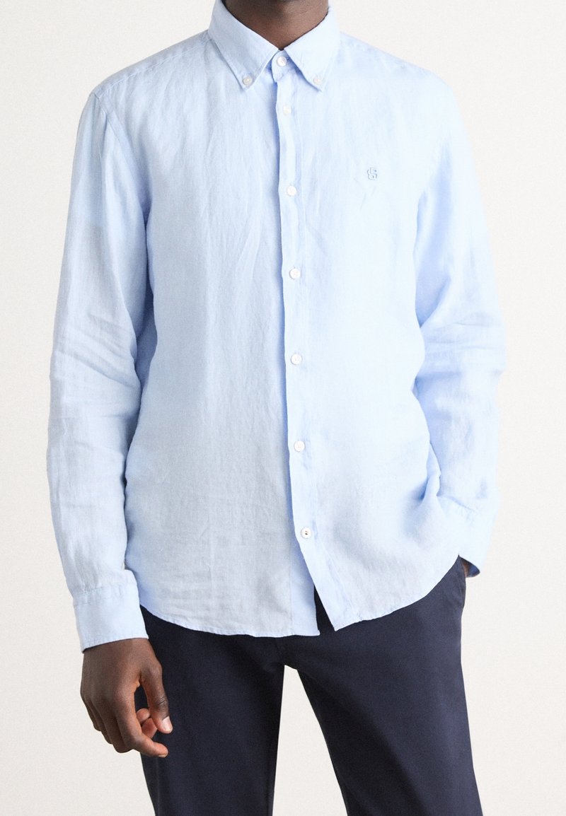 Homme portant une chemise bleu clair à manches longues boutonnée avec col et un pantalon sombre, debout avec une main dans la poche.
