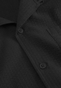 Černá košile z texturované tkaniny s jemným vzorem. Má límec, přední kapsu a černé knoflíky.