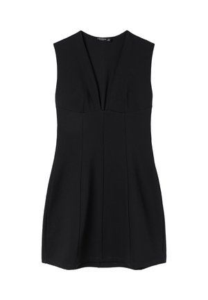 Femme aux cheveux mi-longs portant une robe mini noire moulante sans manches et des escarpins noirs à bout pointu, debout devant un fond gris uni.