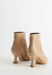 Botines de cuero beige hasta el tobillo con una textura suave, que cuentan con un tacón curvo y un detalle de cremallera dorada en la parte posterior.