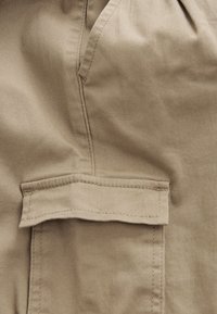 Bézs pamut szövet sima textúrával, téglalap alakú zsebrészlettel és kontrasztos varrással a varrásvonalaknál.