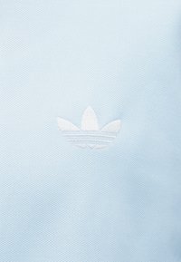 Lyseblå teksturert stoff med en hvit brodert Adidas trefoil-logo plassert midt på flaten.