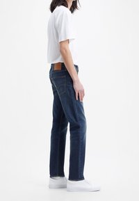 Jeans de mezclilla azul oscuro con pierna recta, ligero desgaste y un parche de cuero marrón en la parte posterior. Combinados con una camiseta blanca y zapatillas blancas.