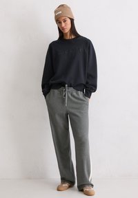 Vrouw in een beige gebreide beanie, een zwarte oversized sweatshirt, grijze joggingsbroek met veters en beige sneakers, staande tegen een witte muur.