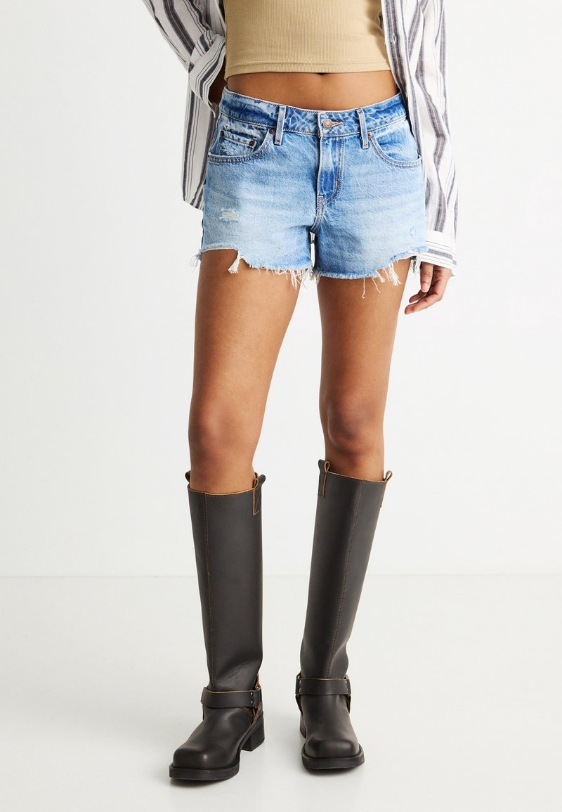 Femme portant un short en jean bleu clair effiloché, un top court côtelé beige, une chemise rayée à manches longues et des bottes en cuir noires montantes jusqu'aux genoux.