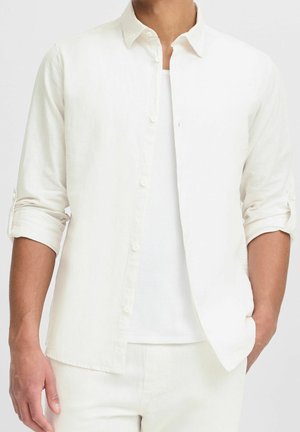 Męski tors ubrany w białą koszulę na guziki z długim rękawem, nałożoną na biały prążkowany podkoszulek, z podwiniętymi rękawami i jedną ręką w kieszeni.